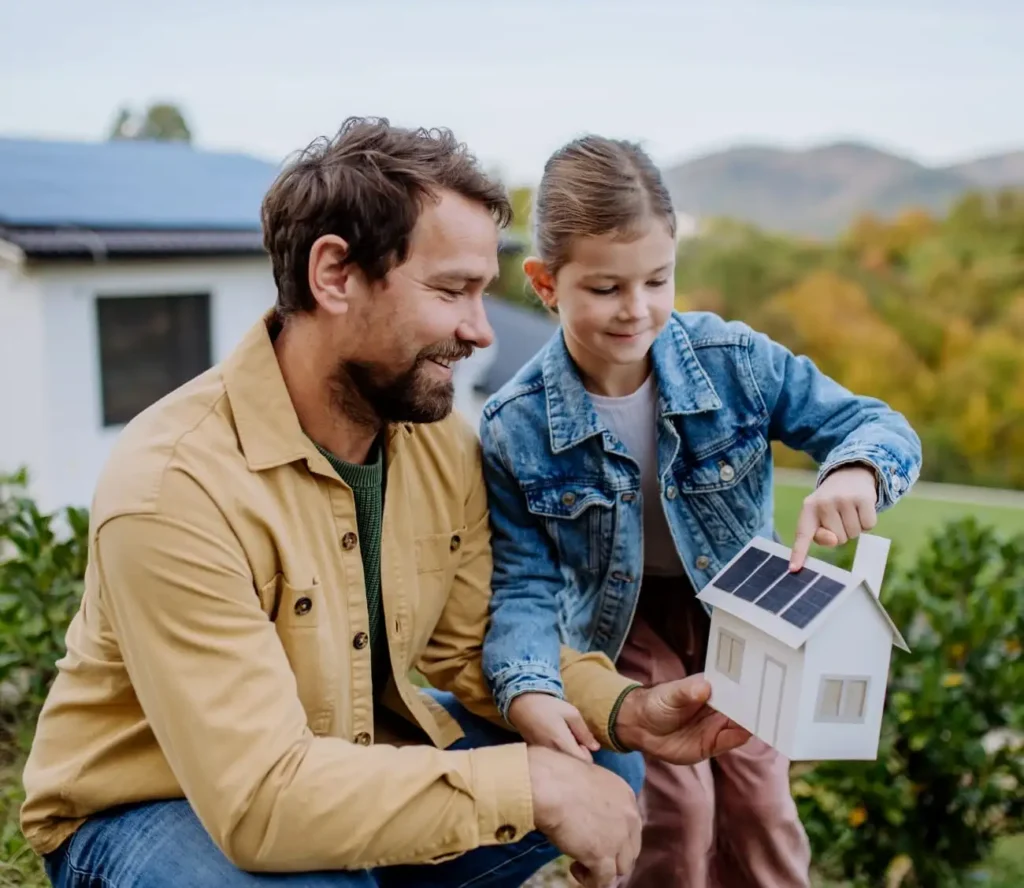 A man and a young girl holding a small model house with a solar panel on its roof, symbolizing clean energy